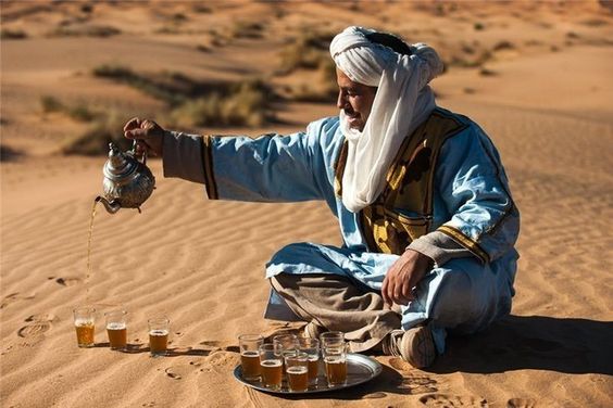 Man in a white turban pouring tea into small glasses on desert sand