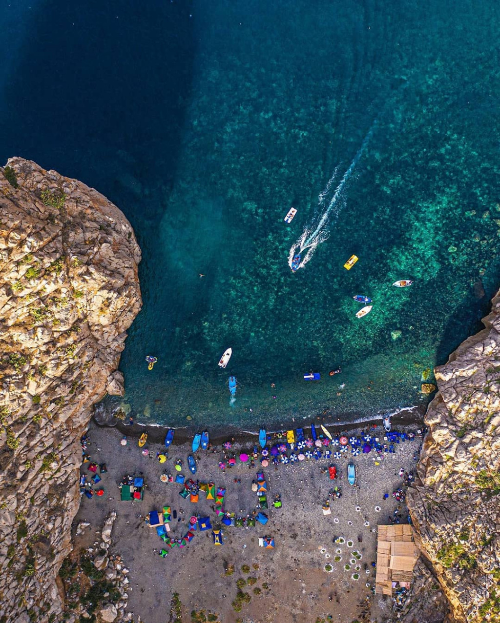 Aerial view of a crowded pebble beach between tall cliffs with boats offshore
