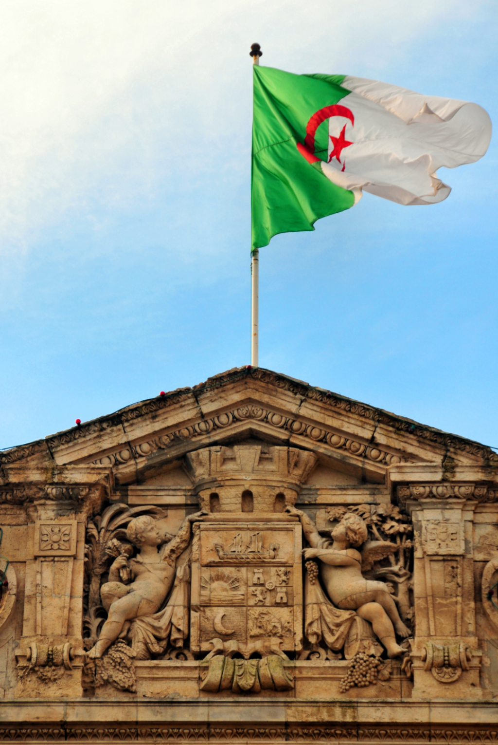 Algerian flag flying above a carved stone pediment with cherubs on a colonial building