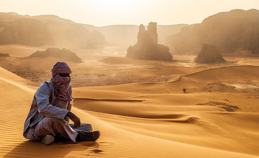 Tuareg man in a turban sitting on golden dunes below sandstone rock towers in the Sahara