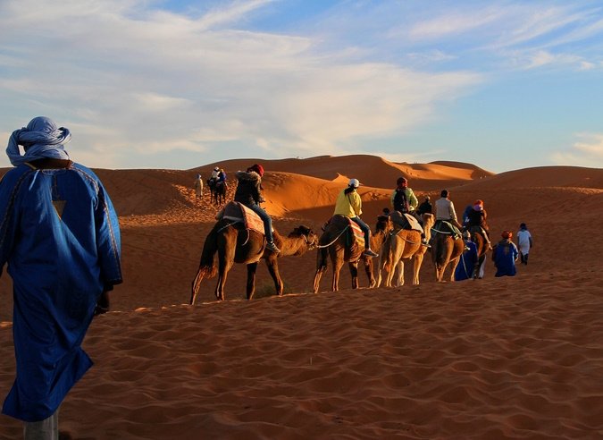 Camel caravan with tourists crossing orange Sahara dunes, led by a guide in blue