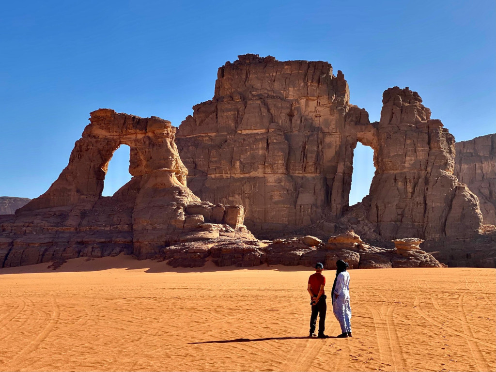 Two people standing on orange sand beneath natural rock arches in the Tassili n'Ajjer