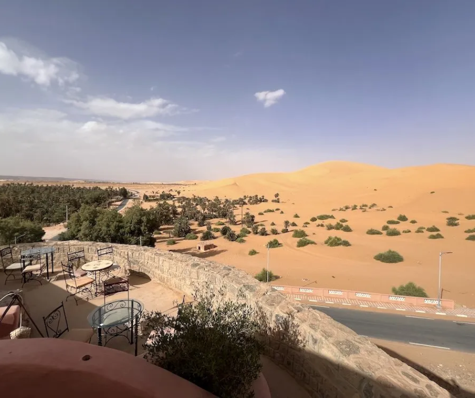 Guesthouse terrace with metal tables overlooking a palm grove and orange dunes in the Algerian Sahara