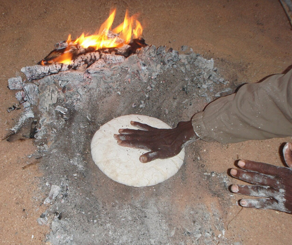 Hand brushing embers away from a round flatbread baking in sand, a taguella, beside a fire