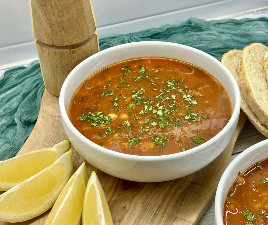 Bowl of red chorba soup with herbs, lemon wedges and bread