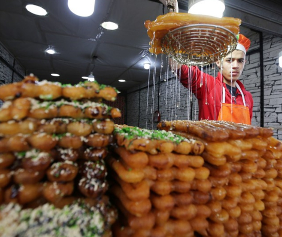 Baker lifting fried zlabia from syrup above stacked piles of sweets