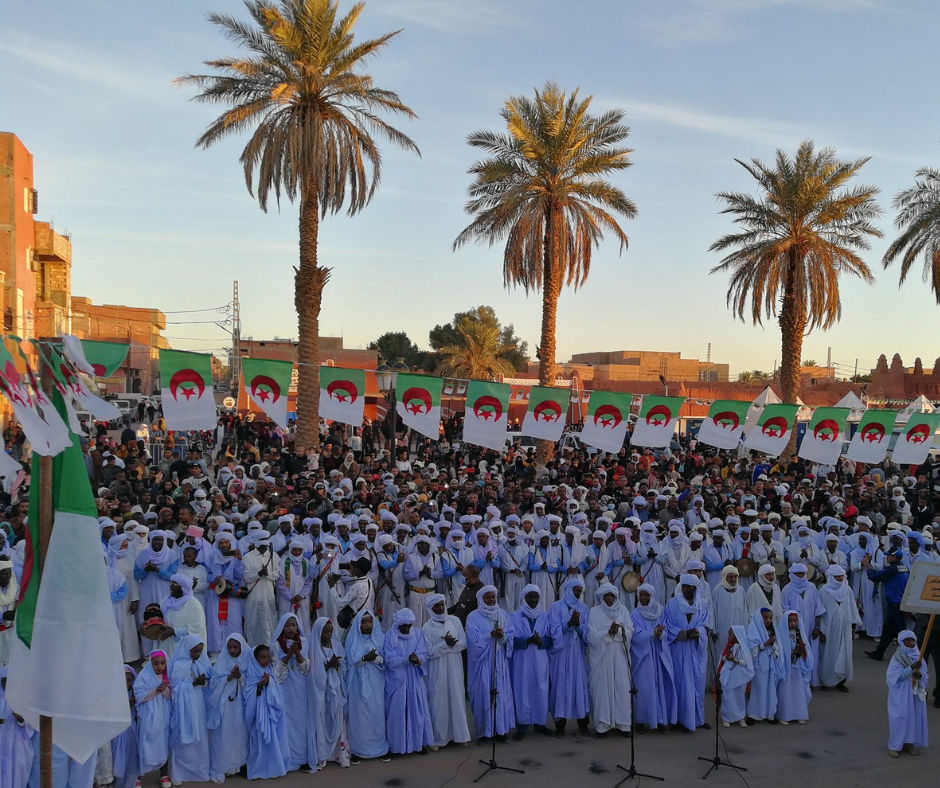 Crowd in blue and white robes under palms with Algerian flags at an Ahellil festival